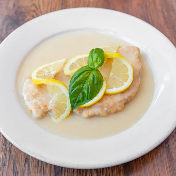 Chicken piccata with lemon slices and a basil leaf, served in a creamy sauce on a white plate.