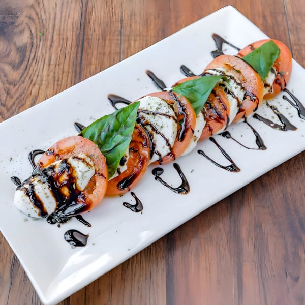 Caprese salad with sliced tomatoes, mozzarella, fresh basil, and balsamic glaze on a white plate.