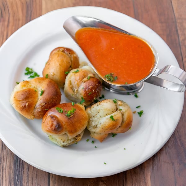 Garlic knots with parsley on a white plate, served with a side of marinara sauce.