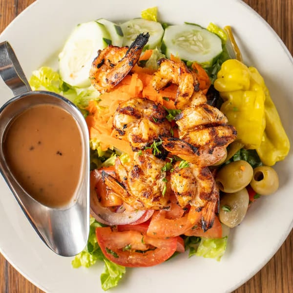 Grilled shrimp salad with lettuce, cucumbers, tomatoes, olives, and a side of dressing.
