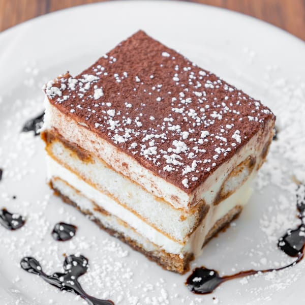 A slice of tiramisu with cocoa powder and powdered sugar on a white plate with chocolate drizzle.