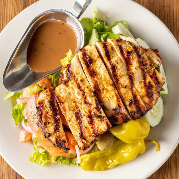 Grilled chicken salad with lettuce, tomatoes, cucumbers, peppers, olives, and a side of dressing.
